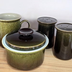 May include: A collection of four ceramic kitchen items in green and brown tones. The set comprises a creamer jug, a lidded casserole dish, and two shakers. The items have a glossy finish and a mid-century modern design.