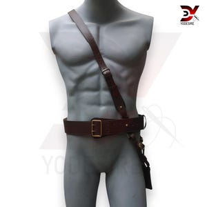 Pu&ograve; includere: Una cintura in pelle marrone con una fibbia dorata e una tracolla in pelle marrone abbinata. La tracolla &egrave; indossata sulla spalla e fissata alla cintura all'altezza del fianco.