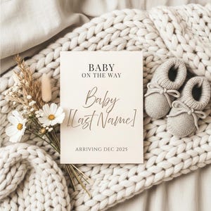 Könnte beinhalten: Eine cremefarbene Schwangerschaftsankündigungskarte mit dem Text "Baby on the Way" und "Baby [Last Name]", mit dem Ankunftsdatum Dezember 2025. Die Karte wird von einem Strauß weißer Blumen und grauen Babyschuhen begleitet.