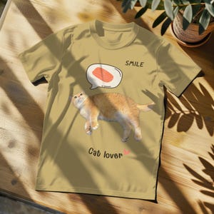 Puede incluir: Una camiseta beige con un gato de dibujos animados y una burbuja de diálogo que dice "SMILE". El texto "Cat lover" está impreso debajo del gato.
