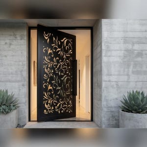 Puede incluir: Una puerta de entrada negra con un intrincado diseño floral cortado en el metal. La puerta está ligeramente entreabierta, revelando un vistazo del interior. La puerta está enmarcada por un borde negro y colocada contra una pared de hormigón. Dos suculentas en macetas flanquean la entrada.