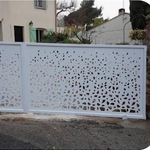 Op de afbeelding: Een witte metalen poort met een bladachtig patroon uitgesneden in de panelen. De poort staat voor een huis met een lichtgekleurde buitenkant en een groene luik. De poort wordt waarschijnlijk gebruikt voor een oprit of tuin entree.