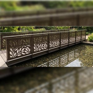 以下が含まれることがあります： 繰り返し葉の模様が施されたレーザーカットパネルが特徴の、ダークブラウンの金属製歩道橋。平らな手すりと垂直の支柱があり、暗い川石が敷き詰められた浅い池の上に架かっています。
