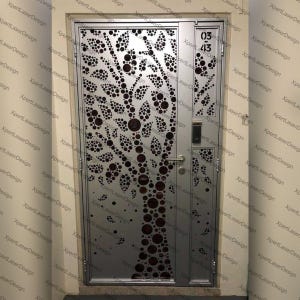 Puede incluir: Una puerta de metal plateado con un diseño de árbol cortado con láser. La puerta tiene un tirador, un teclado pequeño y los números "03" y "43" en negro. La puerta está situada en una entrada de color neutro.