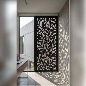 May include: A black metal decorative screen with a leaf pattern is positioned in front of a large window. The screen casts leaf-shaped shadows on the adjacent wall and floor. The room has a modern design with concrete walls and a minimalist desk.