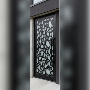 Peut inclure: Une porte en métal noir avec un motif décoratif d'ovales et de cercles qui se chevauchent. La porte est dotée d'une longue poignée noire et de panneaux de verre dépolis derrière la ferronnerie. La porte est encastrée dans un extérieur en béton et gris foncé.