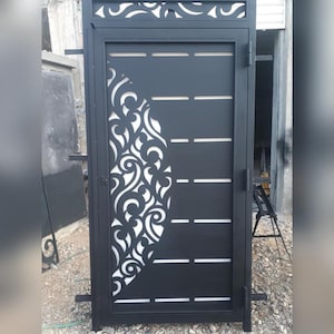 May include: A black metal gate featuring a decorative white swirling design on the right. The gate has horizontal cutouts and a decorative top. The gate is closed and appears to be outdoors.