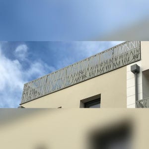 May include: A light grey metal balcony railing featuring a laser-cut pattern of intersecting thin lines and geometric shapes. The railing is mounted on the exterior edge of a beige building roofline against a bright blue sky with white clouds.