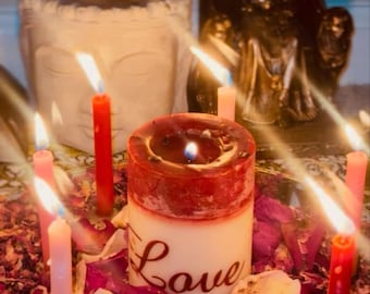 May include: A candlelit display featuring a large, two-toned candle with the word "Love" written on it. Surrounding the central candle are several smaller candles, a heart-shaped candle, and dried rose petals. A statue is in the background.