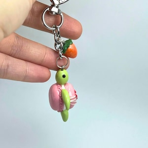May include: A novelty keychain with a green turtle charm and a pink macaron shell. The keychain also features a small orange strawberry charm. The charms are attached to a silver-coloured metal clasp and chain.