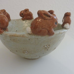 Op de afbeelding: Een keramische kom met een lichtbeige buitenkant en bruine konijnenfiguren rond de rand. De kom heeft een rustieke, handgemaakte uitstraling met kleine bloemendetails aan de buitenkant. De konijnen hebben witte staartaccenten.