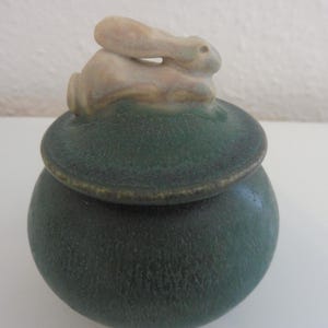 May include: A green ceramic jar with a lid shaped like a white rabbit. The jar has a speckled glaze.