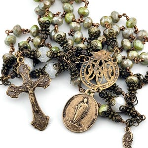 May include: A detailed rosary with olive green and gray faceted beads, connected by antique bronze-colored metal links. The rosary features a cross, a medallion with a religious figure, and a decorative charm. The beads are arranged in a traditional pattern.