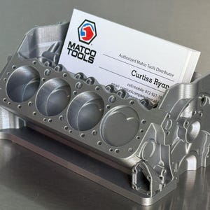 May include: A silver metal engine block shaped business card holder. The holder has a business card inside that reads "MATCO TOOLS" and "Authorized Matco Tools Distributor". The card also has the name "Curtiss Ryan" and contact information.