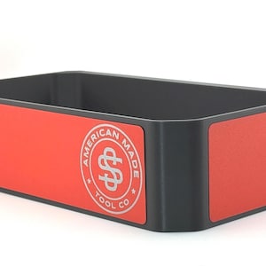 May include: Rectangular black storage bin with red panels. The red panels feature a silver logo that reads "AMERICAN MADE TOOL CO" in a circular design. The bin is empty and sits on a white surface.