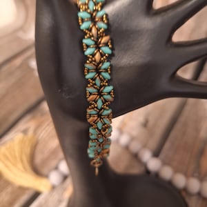 May include: A turquoise and gold beaded bracelet with a geometric pattern. The bracelet is made with small, round beads and is strung on a clear elastic cord.