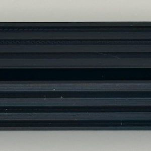 May include: A black plastic 3D printed object with a rectangular shape and a long, thin slot in the centre. The object has a textured surface with raised lines.