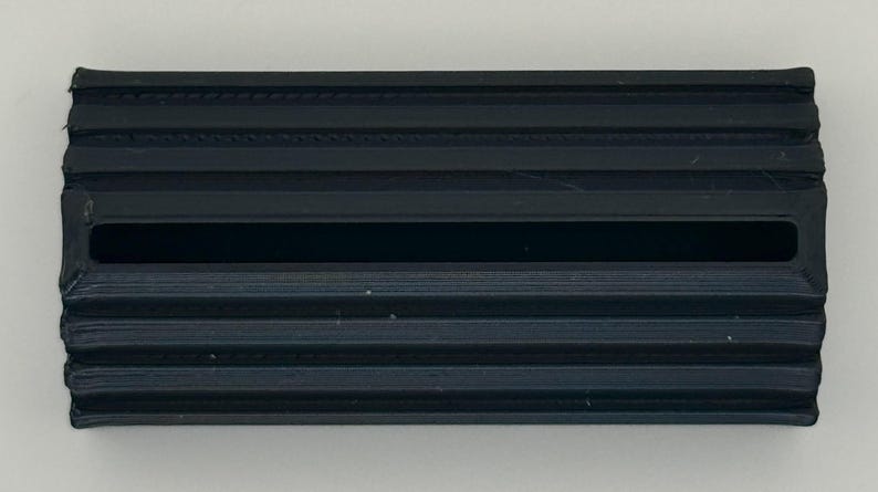 May include: A black plastic 3D printed object with a rectangular shape and a long, thin slot in the centre. The object has a textured surface with raised lines.