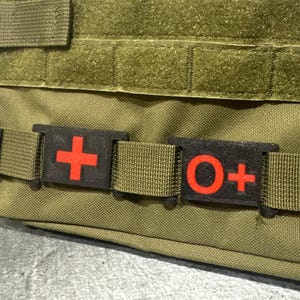 May include: Olive green tactical bag with two black patches. One patch features a red cross, and the other displays the letters "O+" in red. The bag has a woven strap and a "WYNEX TACTICAL" label.