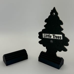 May include: Black car air freshener shaped like a pine tree with the text "Little Trees" and a registered trademark symbol. The air freshener is on a black base.