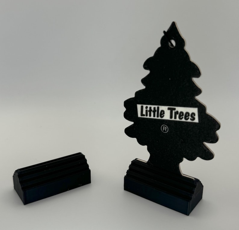 May include: Black car air freshener shaped like a pine tree with the text "Little Trees" and a registered trademark symbol. The air freshener is on a black base.