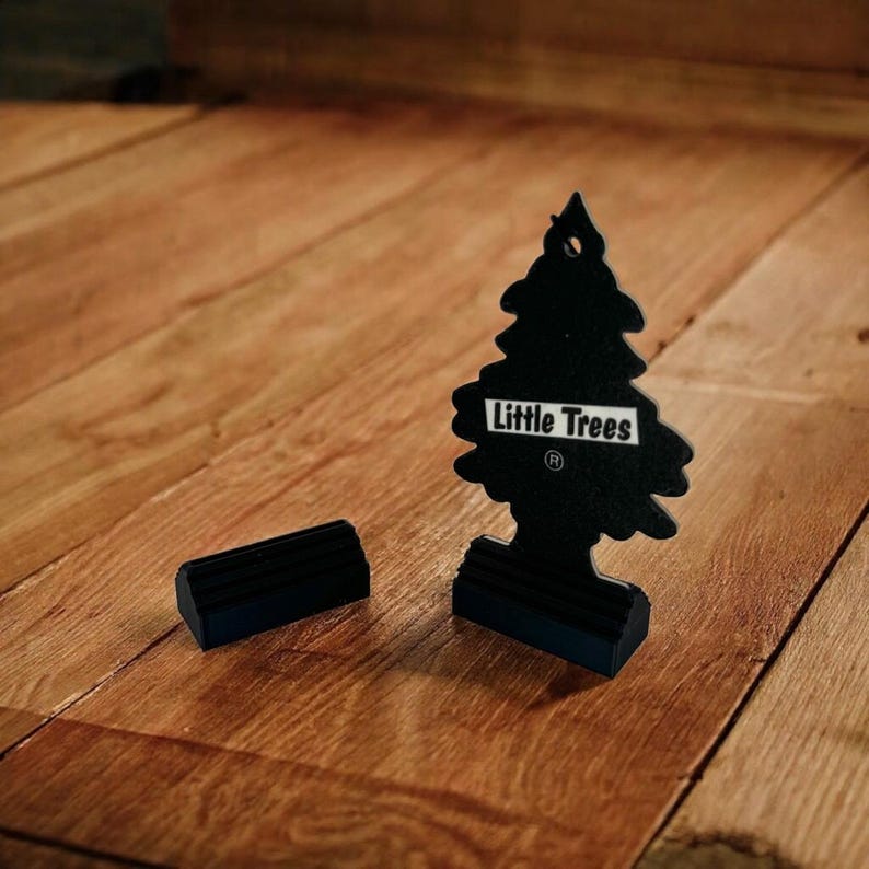 May include: Black car air freshener shaped like a pine tree with the text "Little Trees" on the front. The air freshener is sitting on a wooden surface.