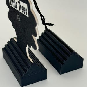 May include: Two black plastic car air freshener holders with a Little Trees car air freshener in the shape of a tree. The air freshener is black with white text that reads "Little Trees" and a registered trademark symbol.