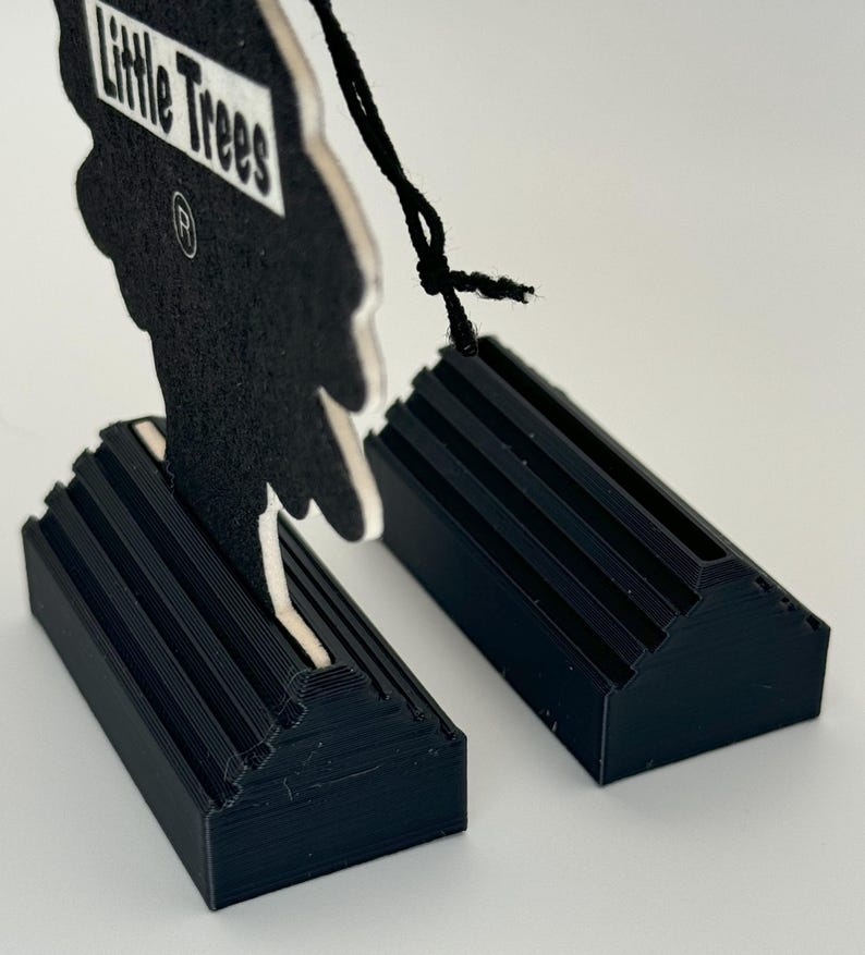 May include: Two black plastic car air freshener holders with a Little Trees car air freshener in the shape of a tree. The air freshener is black with white text that reads "Little Trees" and a registered trademark symbol.