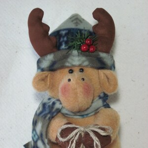May include: A plush brown reindeer wearing a blue and grey patterned hat with a green sprig and red berries. The reindeer is holding a string of bells, including gold, silver, and red.