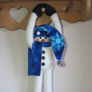 Puede incluir: Un muñeco de nieve blanco con un gorro y una bufanda azules con copos de nieve, colgado de una estantería de madera. El muñeco de nieve tiene ojos negros de botón y una nariz roja. El muñeco lleva zapatos negros con copos de nieve blancos.