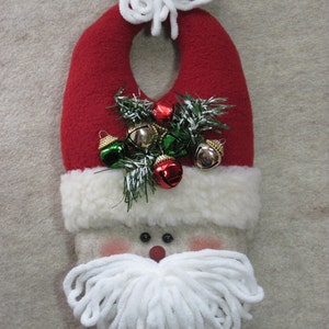 Puede incluir: Una media de Navidad de fieltro roja y blanca con un pompón blanco en la parte superior. La media tiene un borde de piel blanca y una barba de hilo blanco. La media está decorada con cascabeles verdes y rojos.