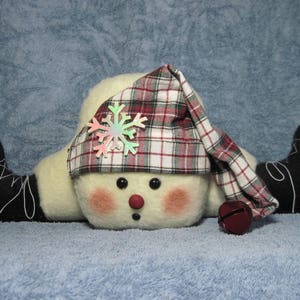 May include: A white snowman with red cheeks and black eyes wearing a red and white plaid hat with a snowflake on top and black boots with snowflakes on them.
