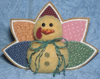 Puede incluir: Un pavo de peluche con un cuerpo marrón, tostado y blanco, una papada roja y un lazo verde. El pavo está sentado sobre un fondo de tela de retazos con un patrón marrón, rosa, verde y azul.