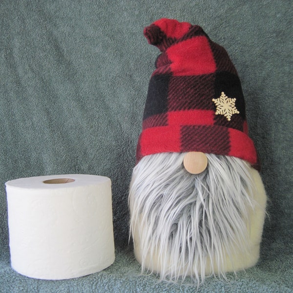 Puede incluir: Un gnomo de cuadros rojos y negros con una barba blanca y esponjosa y una nariz de madera. El gnomo lleva un pin de copo de nieve en su gorro. Un rollo de papel higiénico blanco está en el fondo.