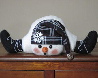 May include: A white snowman with black and red plaid hat, black boots, and a small orange nose. The snowman is lying on its back with its arms outstretched.