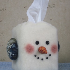 May include: A white, snowman-shaped tissue box cover with black eyes, a carrot nose, and a stitched smile. The snowman has pink cheeks and blue and white earmuffs. White tissues are pulled from the top.