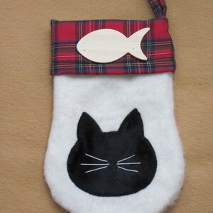May include: A white Christmas stocking with a black cat face and whiskers. The top is red plaid with a wooden fish accent. A red plaid loop is attached for hanging.