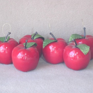 May include: A collection of six glossy red apple ornaments with green leaves and thin wire stems. The apples are evenly spaced and appear to be made of a smooth material, possibly ceramic or plastic.