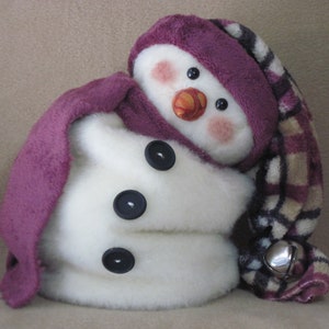 Könnte beinhalten: Ein weißer Plüsch-Schneemann, der einen lila-braunen karierten Schal und Hut mit einer Glocke trägt. Der Schneemann hat schwarze Knopfaugen und eine Karottennase.