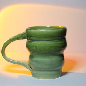 May include: A green ceramic mug with a unique, wavy design. The mug has a wide, rounded handle and a smooth, glazed finish.