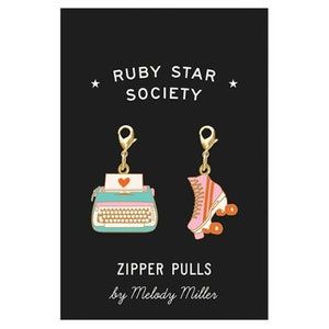 May include: A set of two gold zipper pulls featuring a typewriter and roller skate design. The zipper pulls are part of the Ruby Star Society collection by Melody Miller.