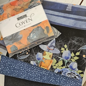 May include: A stack of fabric featuring a Moda Coven by Robin Pickens charm pack in a plastic sleeve, layered with folded fabric pieces. The patterns include orange and black floral prints, a witch illustration, blue polka dots, and solid black.