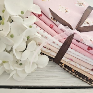 May include: A stack of fabric squares tied with a brown ribbon and a sheer pink ribbon. The fabrics feature various pink patterns, including hearts and teddy bears. White flowers are in the foreground, with a white wooden surface in the background.