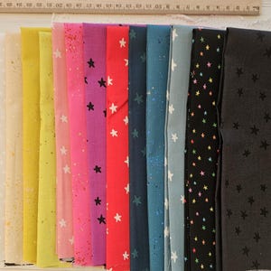 May include: A collection of colorful fabric swatches, each featuring a star pattern. The fabrics vary in color, including white, yellow, pink, red, and black. A wooden ruler is placed at the top of the fabrics.