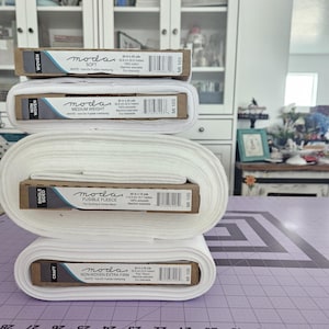 May include: A stack of white fabric rolls and boxes. The boxes are labeled "moda" with descriptions like "Soft," "Medium Weight," and "Fusible Fleece." The rolls vary in size, with the largest labeled "Fusible Fleece" and measuring 45 inches by 13.7 yards.