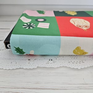 May include: A folded piece of fabric featuring a grid pattern with colorful squares in pink, green, red, light blue, and white. The print includes various illustrations such as a white owl, green holly leaves, and assorted small geometric shapes.