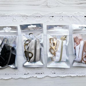 May include: Four clear plastic bags with zippers, each containing a different type of zipper slider and tape. The sliders are gold, silver, and black. The tapes are white, black, and a light brown color.