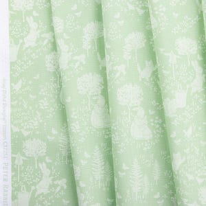 May include: Light green fabric with a repeating pattern of white rabbits, trees, and other foliage. The fabric is folded, showing the texture and design. The text "Riley Blake Designs" and "Peter Rabbit" is visible on the edge.
