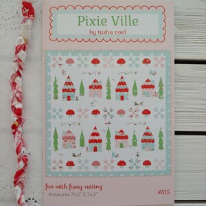 May include: A pink and blue quilt pattern with the title "Pixie Ville" by Tasha Noel. The pattern features a whimsical village with houses, trees, and mushrooms. The pattern measures 74.5 inches by 74.5 inches.