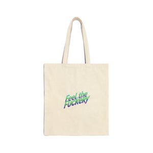 May include: Cream-colored tote bag with the phrase "Feel the Fuckery" in blue and green lettering. The bag has two handles and is made of a canvas-like material. The design is centered on the front of the bag.